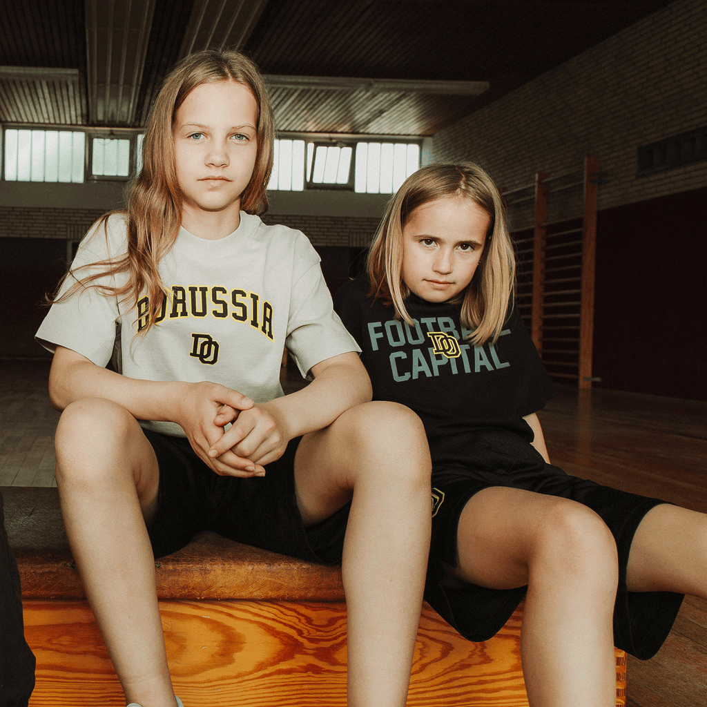 BVB-College-T-Shirt für Kinder, präsentiert von zwei sitzenden Kindern in einem Innenraum. Schwarzes und weißes T-Shirt mit Borussia Dortmund Schriftzug.