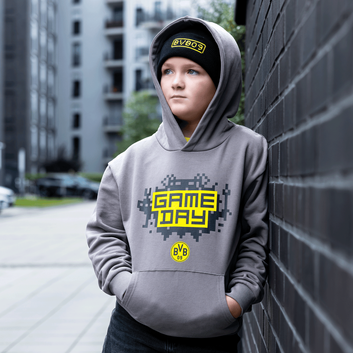 Felpa con cappuccio grigia da gaming del BVB per bambini con stampa “Game Day” in stile pixel e logo del BVB, indossata da un bambino appoggiato a una parete
