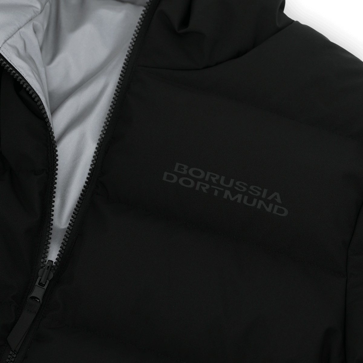 Vue détaillée de la veste réversible du BVB, de couleur noire, dotée d'une surface réfléchissante et du logo du Borussia Dortmund sur la poitrine.