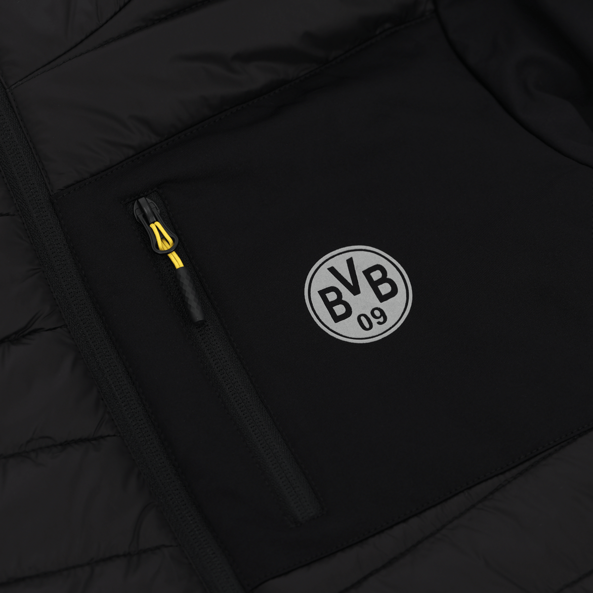 Gros plan sur la veste de stade du BVB 2025, de couleur noire, avec le logo « BVB 09 » blanc et un tirette de fermeture éclair jaune.