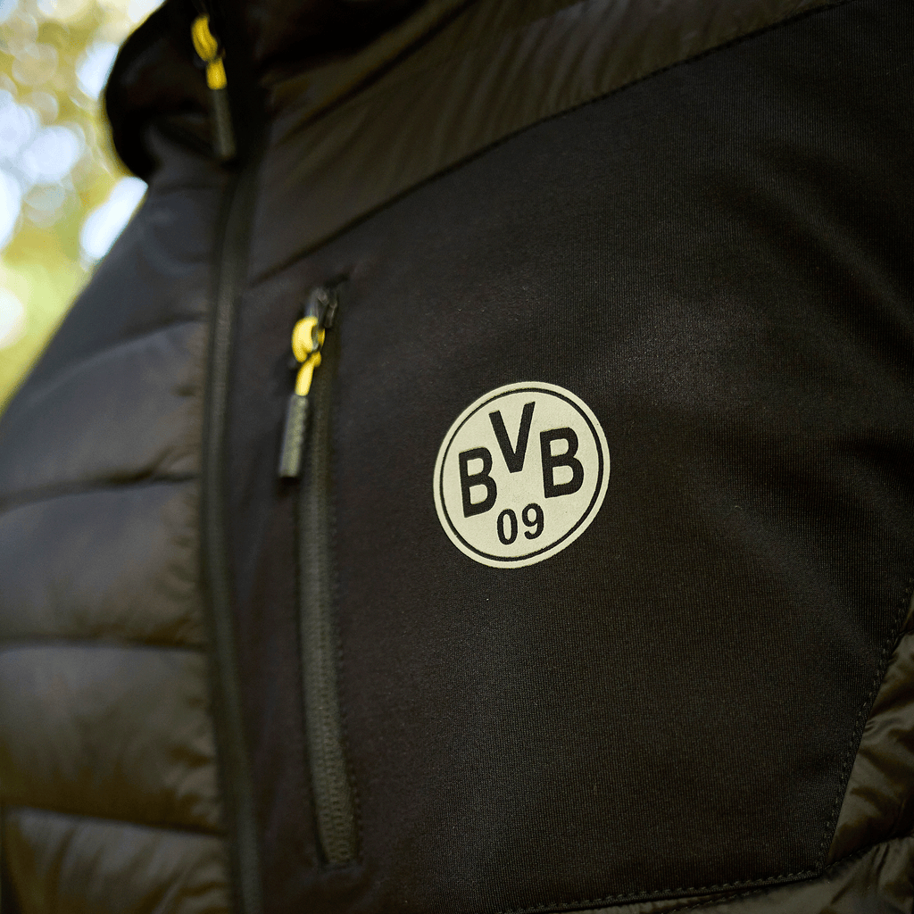 Veste de stade noire du BVB 2025, avec le logo du BVB sur la poitrine et une fermeture éclair jaune, idéale pour les supporters du Borussia Dortmund.