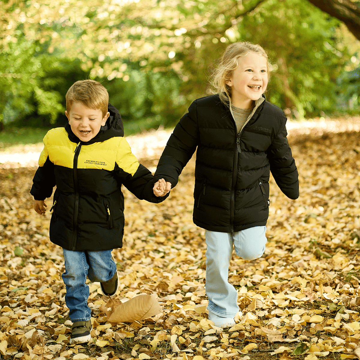 Zwei Kinder, eines in einer schwarzgelben BVB-Wendejacke, das andere in einer schwarzen Jacke, laufen gemeinsam durch herbstliches Laub.
