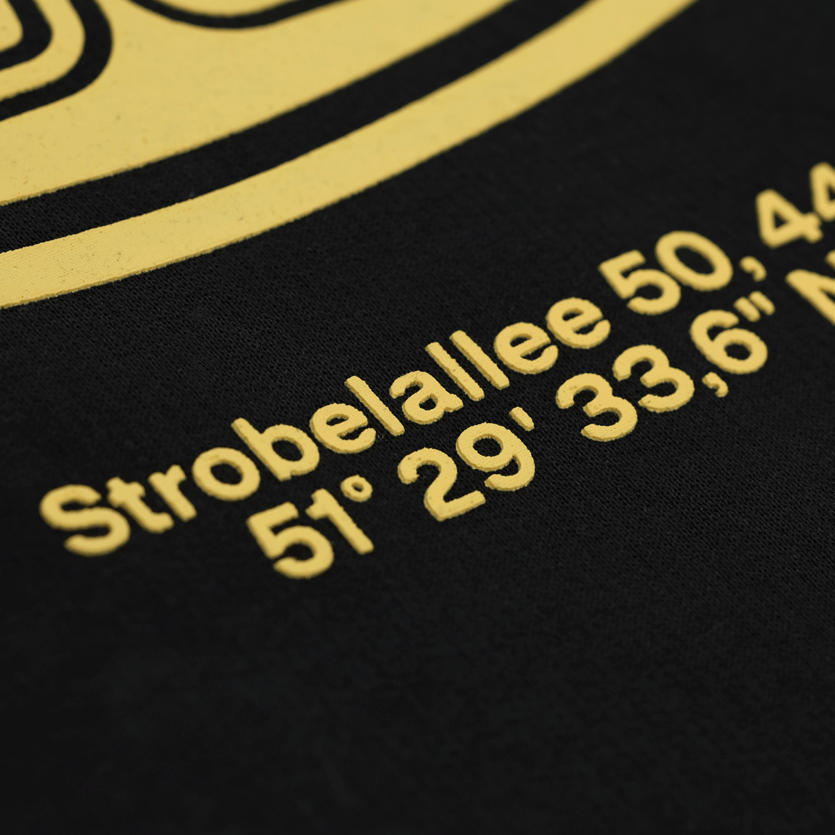 Gros plan sur un sweat à capuche noir du BVB orné d'une inscription jaune « Strobelallee 50 » et des coordonnées 51° 29' 33,6" N. Produits dérivés du Borussia Dortmund.
