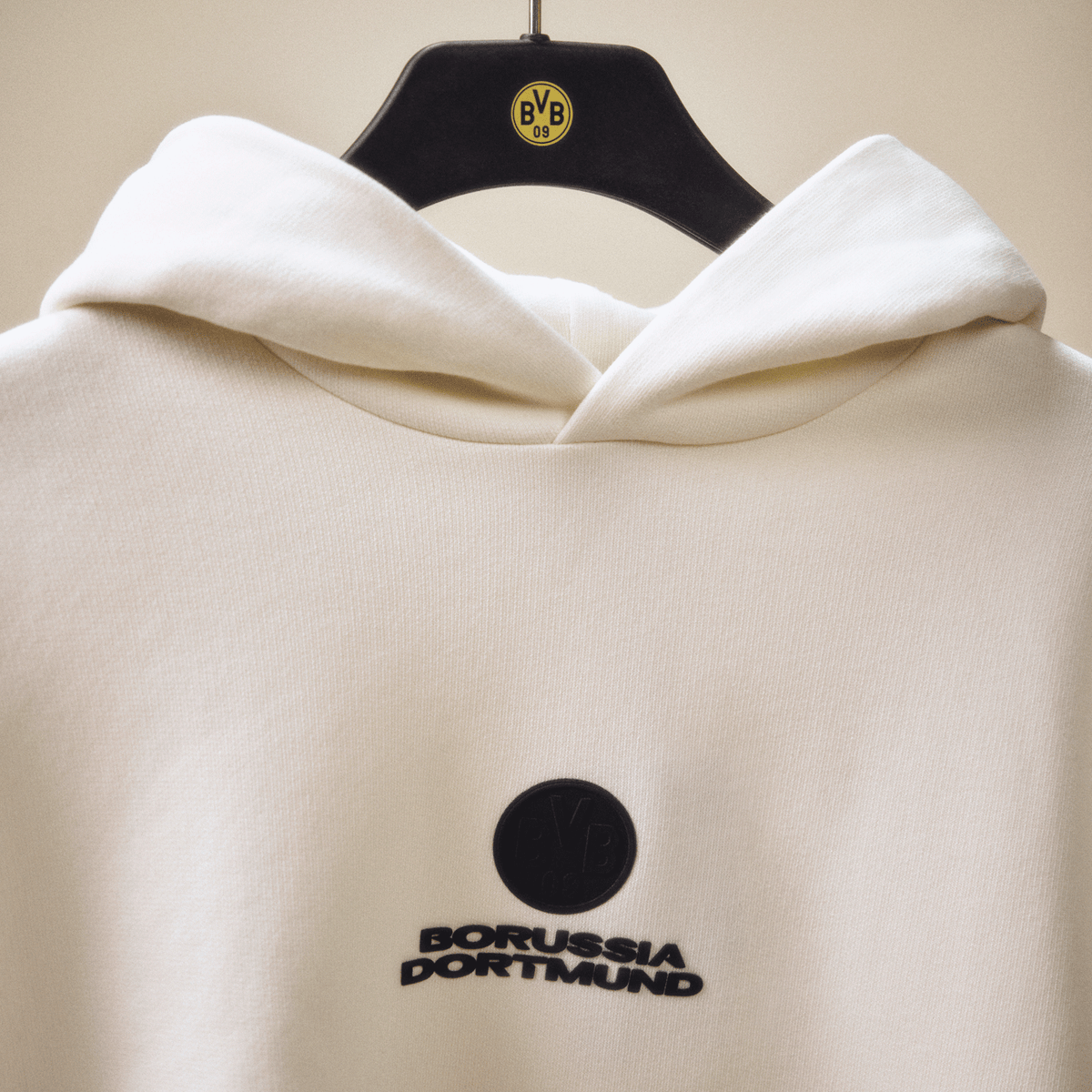 オフホワイトのBVB Coreフーディ。ブラックのボルシア・ドルトムントのロゴが入っており、ハンガーにかけられて展示されています。
