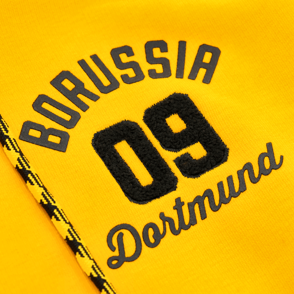 Close-up of the yellow BVB Explorer I sweatshirt jacket featuring an embroidered Borussia Dortmund 09 logo and black-and-yellow decorative patterns.