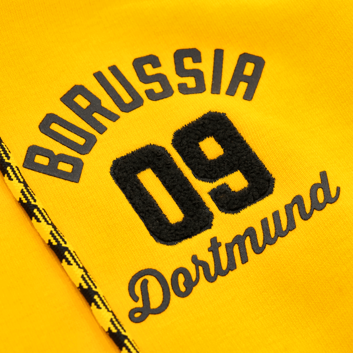 Close-up of the yellow BVB Explorer I sweatshirt jacket featuring an embroidered Borussia Dortmund 09 logo and black-and-yellow decorative patterns.