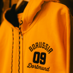Yellow BVB Explorer I sweatshirt jacket with black Borussia Dortmund lettering and 09 logo, hood, and black-and-yellow drawstring details—sporty and comfortable.