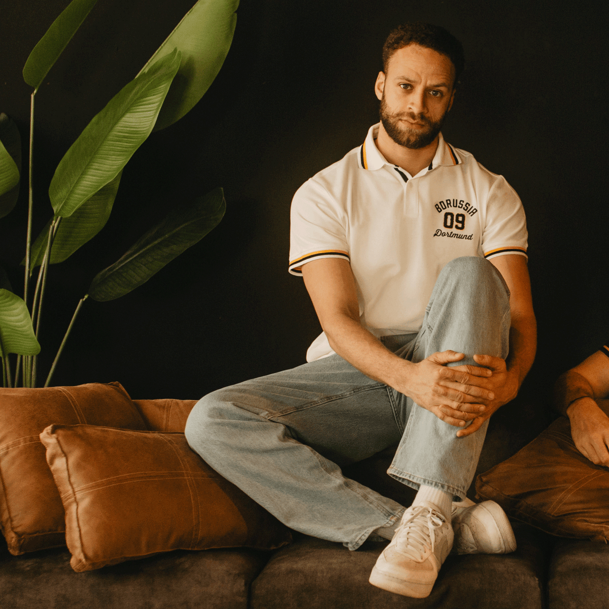 Polo blanc BVB Explorer I avec l'inscription « Borussia Dortmund », idéal pour les supporters. Modèle décontracté, présenté par un mannequin dans une pose détendue, allongé sur des coussins marron.