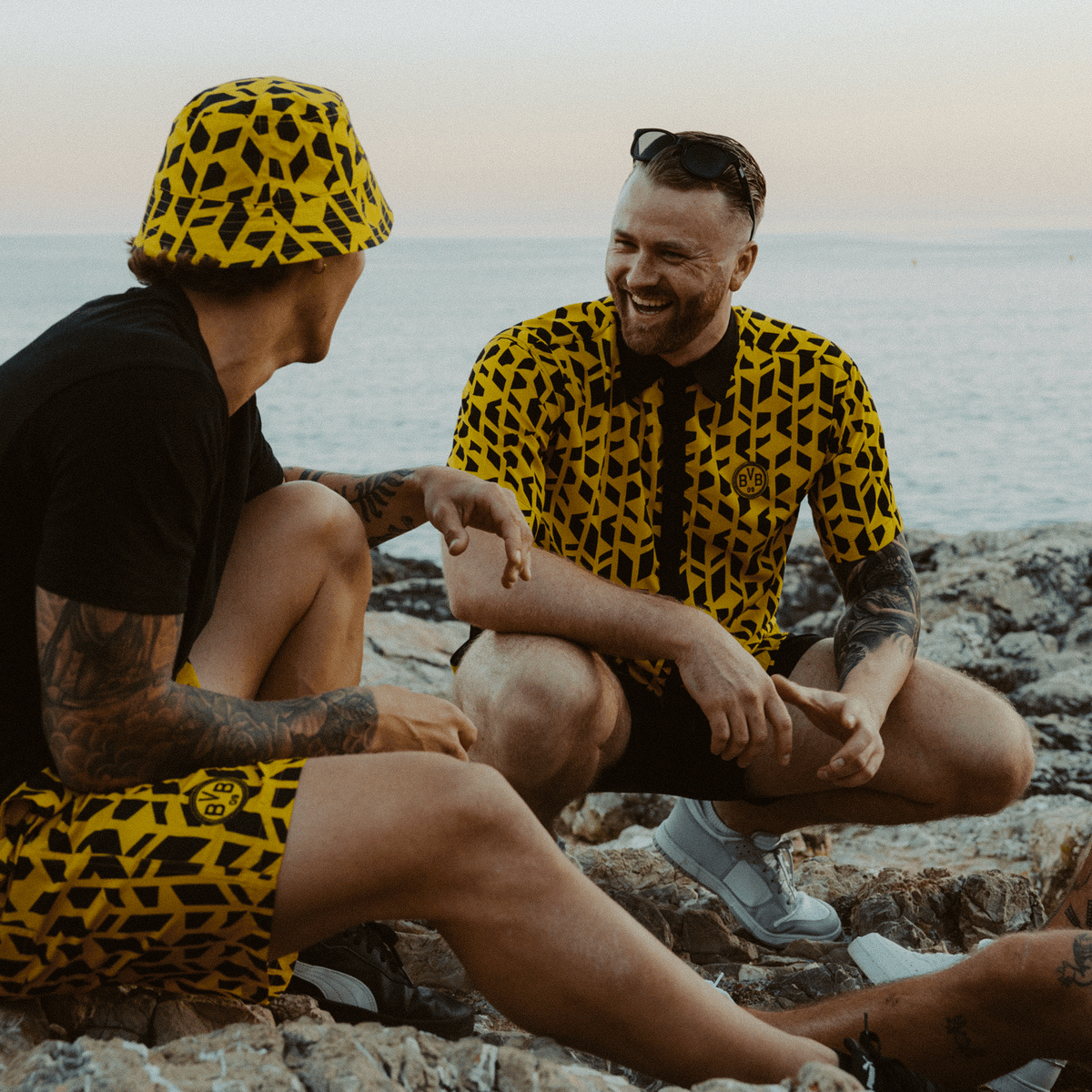 Due uomini indossano la collezione “BVB Festival” nei colori nero e giallo, che comprende una camicia a fantasia e un cappello da pescatore, mentre sono seduti su una spiaggia rocciosa in un’atmosfera estiva.