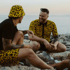 Zwei Männer tragen die schwarzgelbe BVB-Festival-Kollektion, einschließlich gemustertem Hemd und Fischerhut, während sie an einem felsigen Strand in sommerlicher Atmosphäre sitzen.