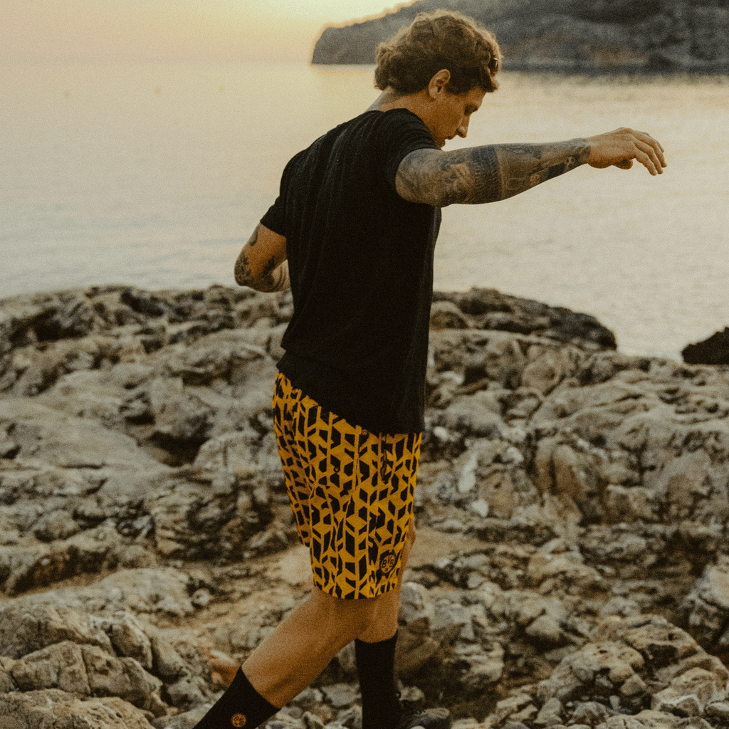 Una persona indossa i pantaloncini del BVB Festival II con il motivo nero e giallo, fotografata in un pittoresco paesaggio roccioso al tramonto.