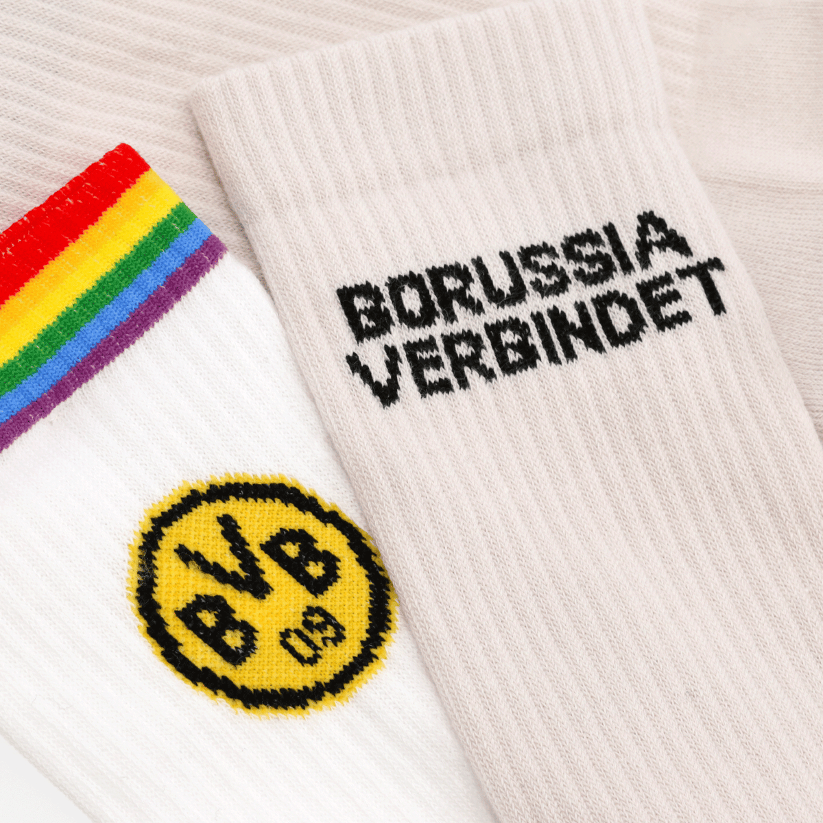Ein Set aus zwei BVB-Socken, eine mit Regenbogenstreifen und gelbem BVB-Logo, die andere in Beige mit dem schwarzen Schriftzug 'Borussia verbindet'.