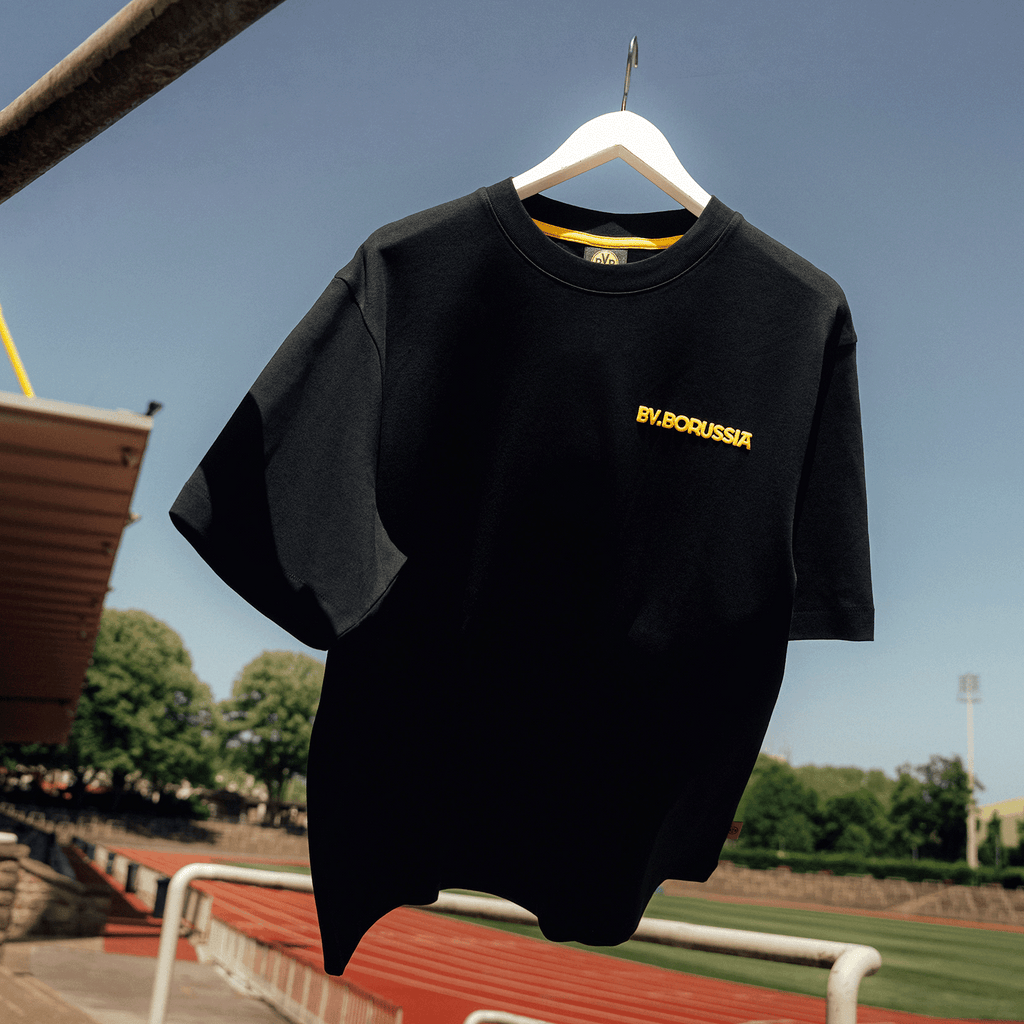 一件黑色多特蒙德（BVB）T恤，背景为红色泥土色，印有黄色“BV.BORUSSIA”字样，悬挂在球场外的一根衣架上。