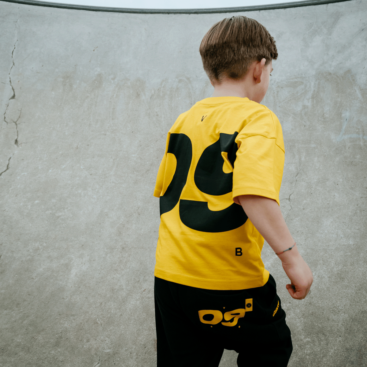 子供用ブラック＆イエローのBVBスウェットパンツ「Urban 09」。黄色のTシャツと合わせ、コンクリートの背景を背にした後ろ姿。