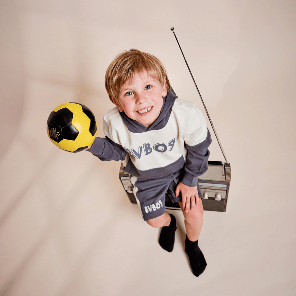 Un bambino indossa la felpa con cappuccio BVB Colorblock II nei colori grigio e bianco con il logo BVB09 e tiene in mano un pallone da calcio nero e giallo. Un articolo perfetto per i piccoli tifosi del BVB.