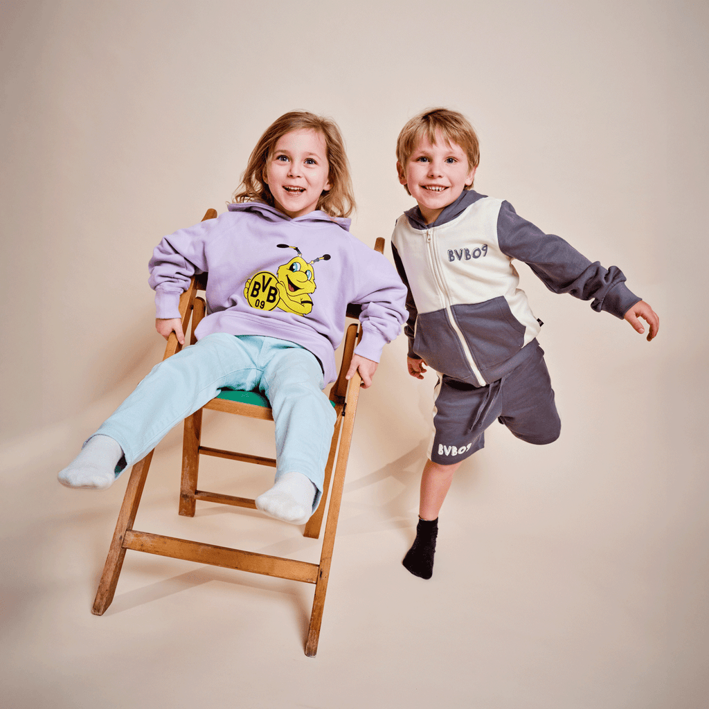 Zwei Kinder präsentieren BVB-Merchandise: Mädchen trägt einen lila Hoodie mit EMMA-Motiv, Junge in grauem BVB 09 Trainingsanzug.