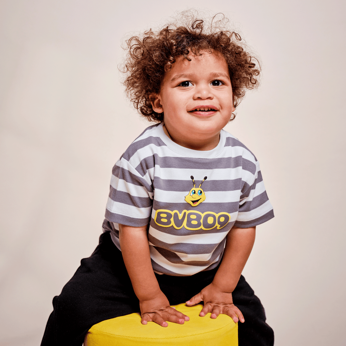 A gray-and-white striped BVB T-shirt for toddlers featuring the EMMA logo and "BVB09" print, modeled by a baby sitting on a yellow stool.