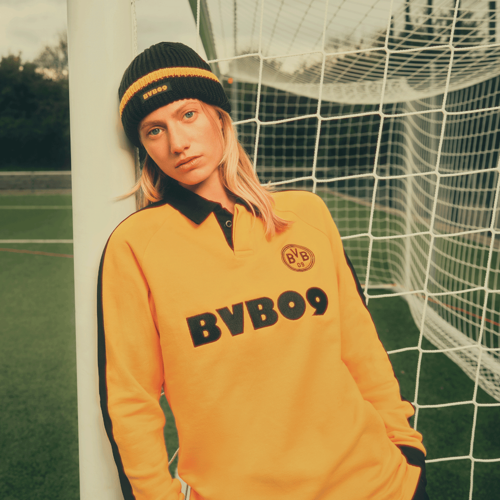 Bonnet noir et jaune du BVB au look rétro des années 70, mis en valeur avec élégance sur un terrain de football avec un but en arrière-plan.