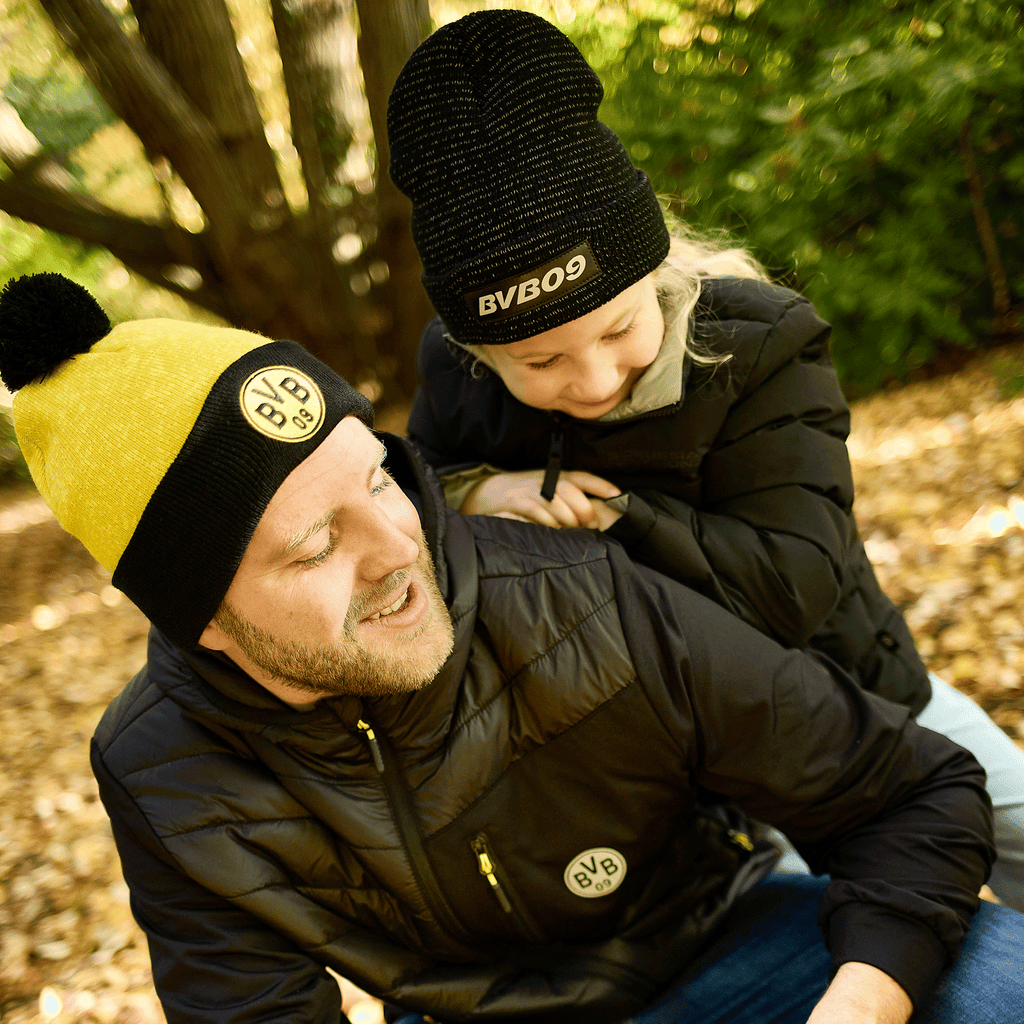 Cappelli con pompon del BVB nei colori nero e giallo e in nero con il logo del BVB, indossati da un uomo e da un bambino in uno scenario autunnale.