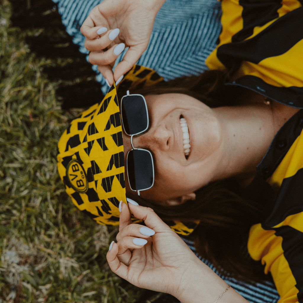 Cappello da pescatore nero e giallo del BVB con motivo geometrico e logo del BVB, indossato da una donna rilassata sdraiata su una coperta.