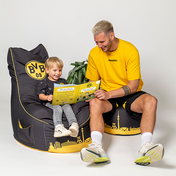 Ein Kind in schwarzgelbem BVB-Outfit sitzt auf einem BVB-Sitzsack und liest gemeinsam mit einem Erwachsenen das BVB-Mitmachbuch.