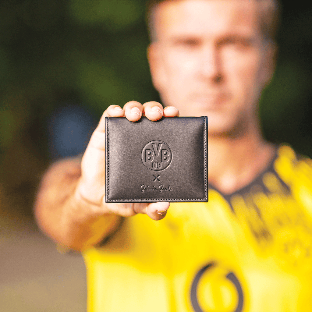 Schwarze Leder-Geldbörse von BVB x Jaimie Jacobs mit geprägtem BVB 09-Logo, gehalten in der Hand eines Mannes vor unscharfem Hintergrund.