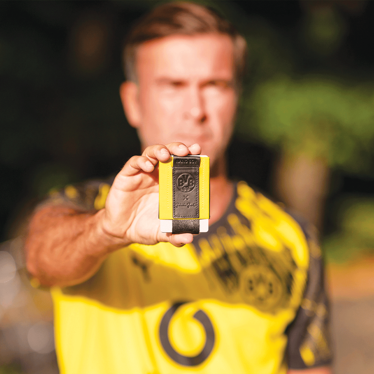 Porte-monnaie BVB Nano Boy Pocket aux couleurs noir et jaune, maniable et pratique, avec en arrière-plan une personne portant un maillot du BVB.