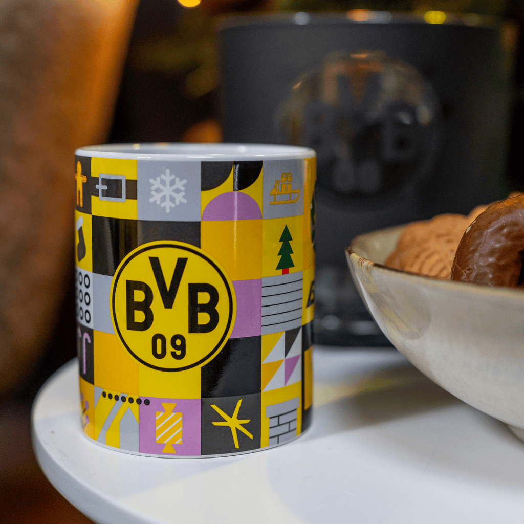 Das Bild zeigt eine farbenfrohe BVB-Winter-Weihnachtstasse im schwarzgelben Design mit BVB-Logo und weihnachtlichen Symbolen, dekorativ auf einem Tisch arrangiert.