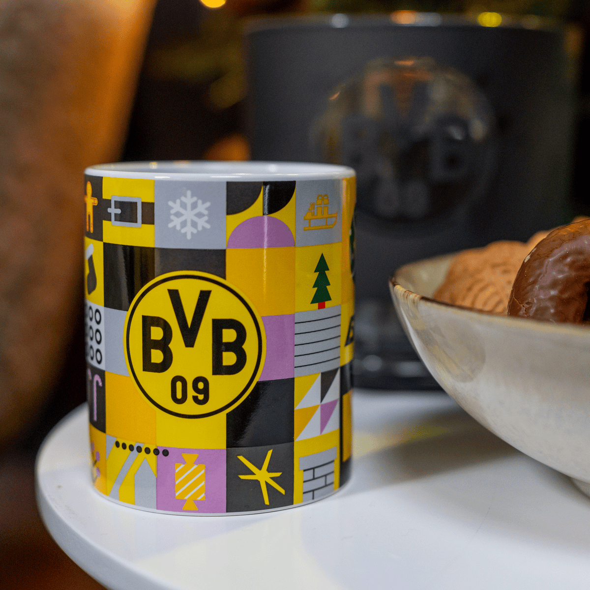 Das Bild zeigt eine farbenfrohe BVB-Winter-Weihnachtstasse im schwarzgelben Design mit BVB-Logo und weihnachtlichen Symbolen, dekorativ auf einem Tisch arrangiert.