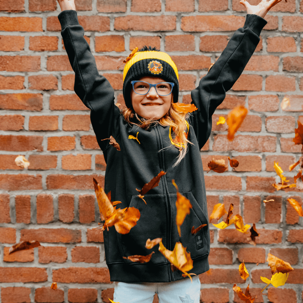 A child is wearing a black-and-yellow BVB children's sweatshirt, set against an autumn scene featuring a brick wall and falling leaves.