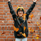 Black-and-yellow BVB Sesame Street hat for children, worn by a smiling child, in an autumnal setting with falling leaves in front of a brick wall.