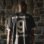 Vue arrière du maillot spécial du BVB « 100 ans de Rote Erde », de couleur noire, avec le numéro 9 doré et l'inscription « BV. Borussia », porté par une personne devant un mur de pierre.