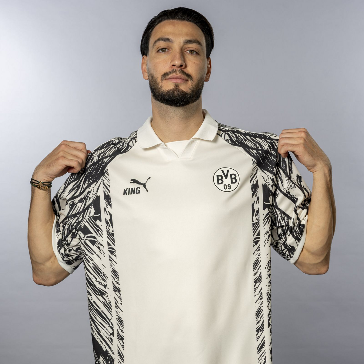La maglia BVB King bianca e nera con motivo astratto in bianco e nero, logo PUMA nero ed emblema del BVB, indossata da un uomo vista di fronte.