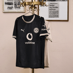 Das schwarze BVB-Sondertrikot 100 Jahre Rote Erde mit goldenen Details, Puma-Logo, BVB-Emblem, Vodafone-Logo und Polestar-Aufdruck, aufgehängt im Vintage-Stil.