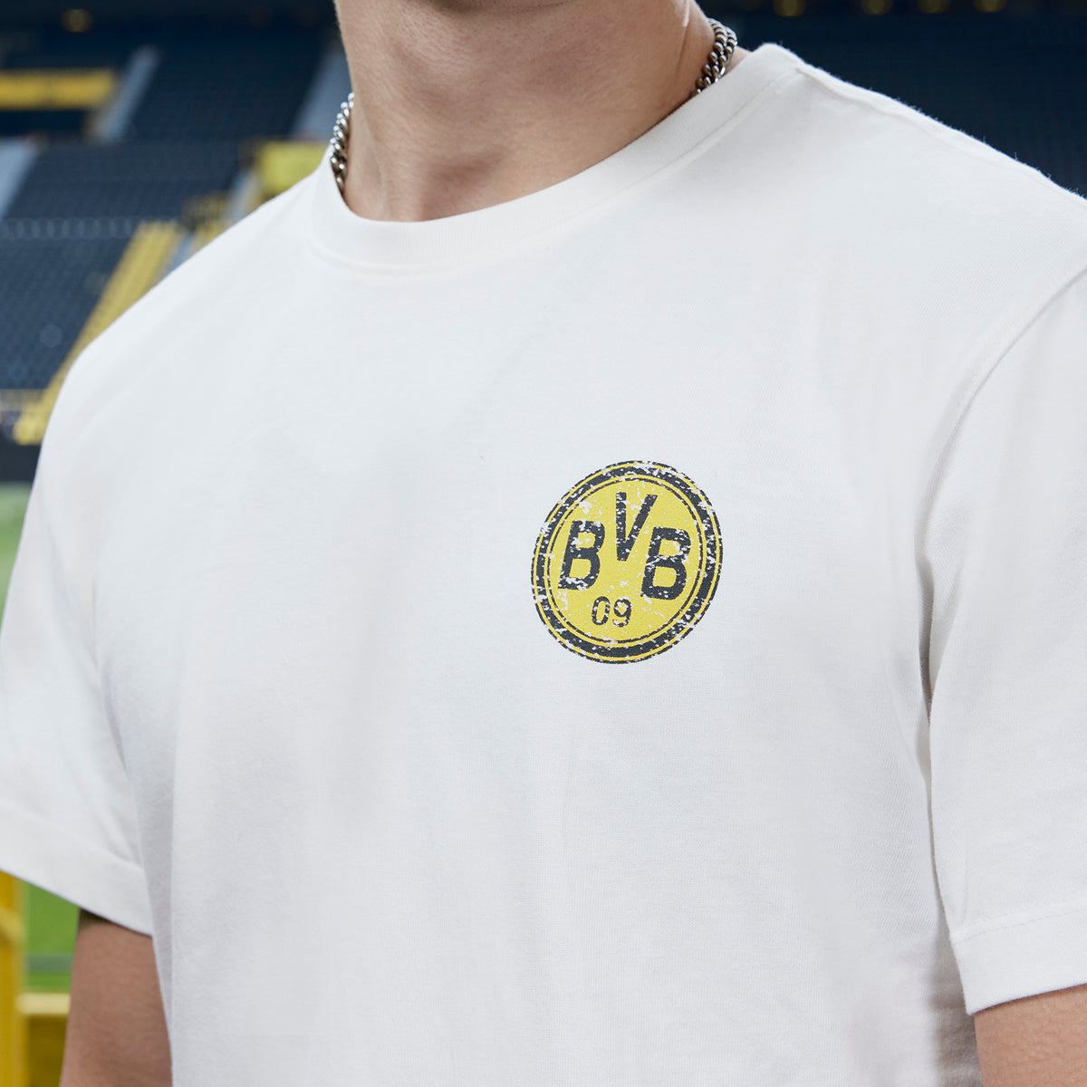 Weißes BVB x Guess T-Shirt mit kleinem BVB-Logo auf der linken Brust, aufgenommen vor der Kulisse des SIGNAL IDUNA PARKs.