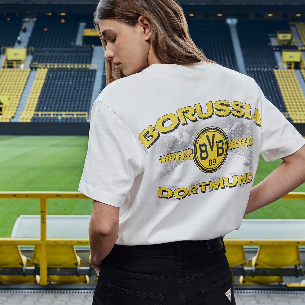 White BVB x Guess T-shirt with a large BVB 09 logo and "Borussia Dortmund" lettering on the back. Worn by a woman at SIGNAL IDUNA PARK.