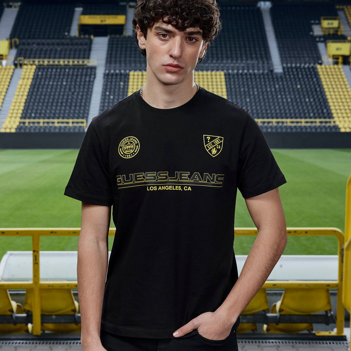 Schwarzes BVB x Guess T-Shirt mit goldfarbenem Guess Jeans Schriftzug und BVB-Emblem, präsentiert im SIGNAL IDUNA PARK.