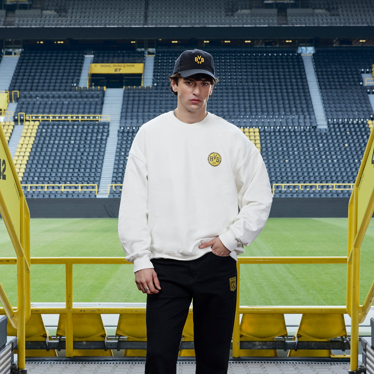 Weißes BVB x Guess Sweatshirt mit gelbem BVB-Logo auf der Brust, getragen von einem Modell im SIGNAL IDUNA PARK mit Fan-Tribünen im Hintergrund.