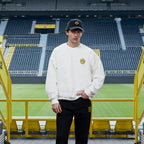 Sweat-shirt blanc BVB x Guess avec le logo BVB jaune sur la poitrine, porté par un mannequin au SIGNAL IDUNA PARK, avec les tribunes des supporters en arrière-plan.