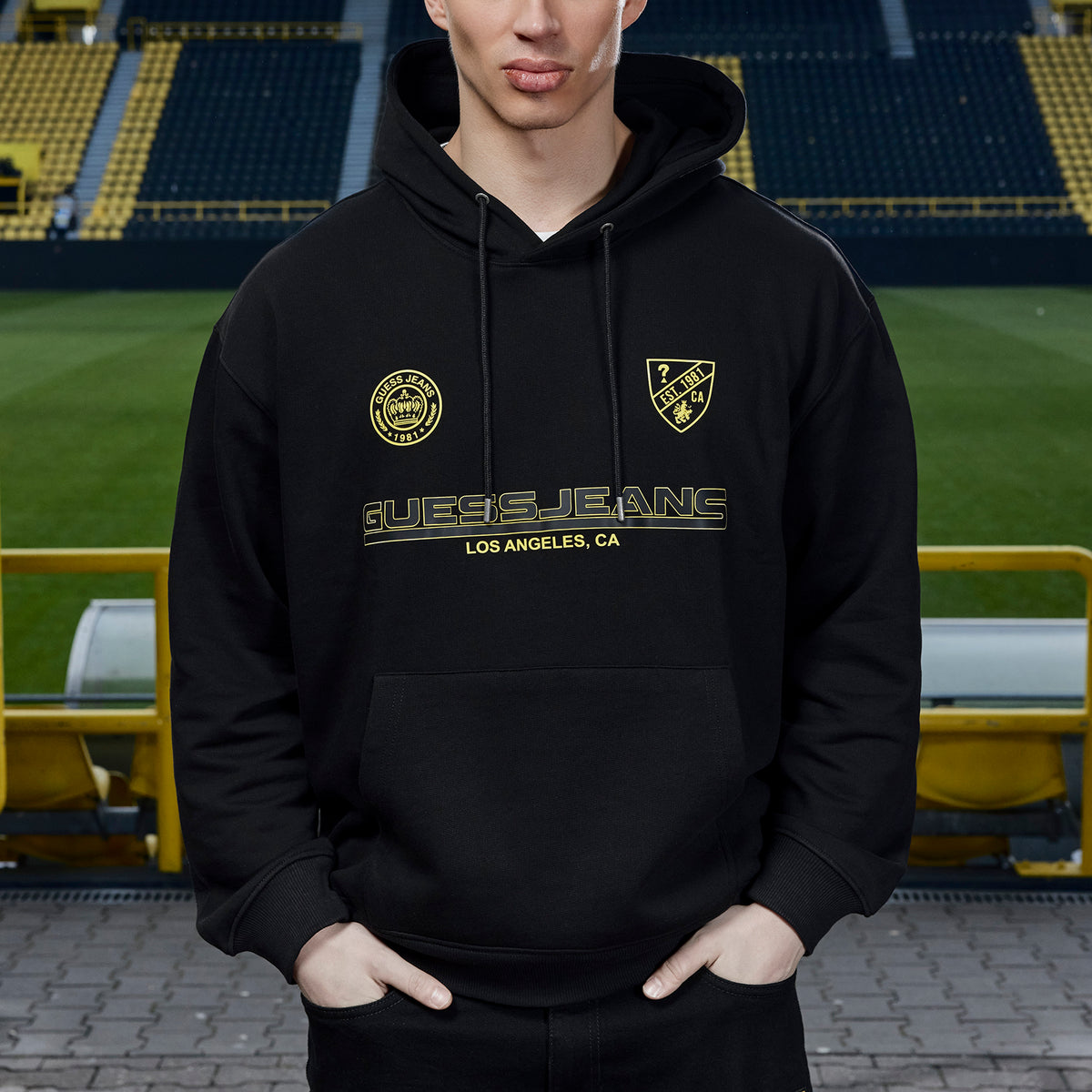 Schwarzer BVB x Guess Hoodie mit gelbem Guess Jeans Logo und Borussia Dortmund Emblemen, präsentiert vor Stadionkulisse, Vorderansicht.