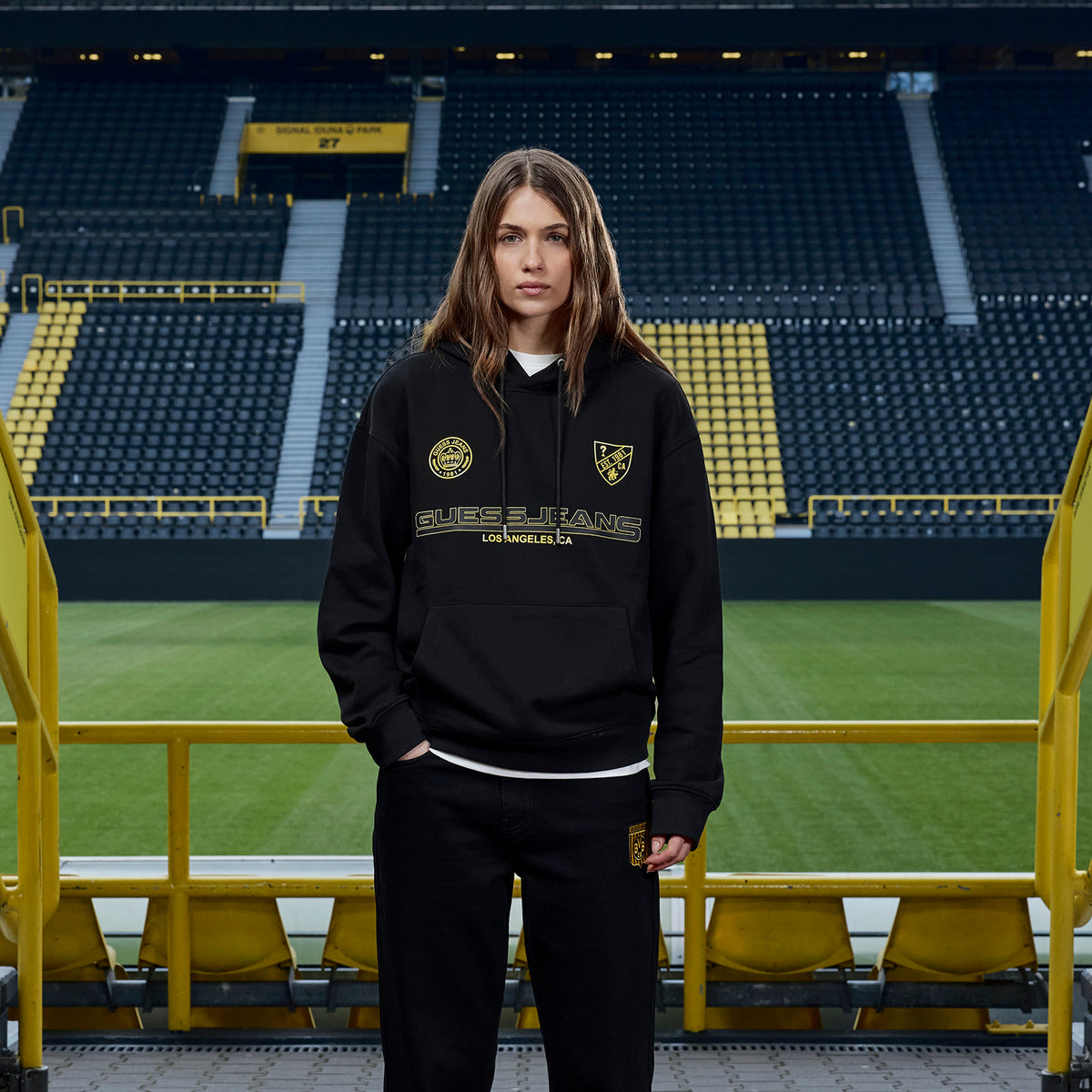 女性が、BVBのロゴと「Guess」の文字が入った黒のBVB × Guessのフーディーを、シグナル・イドゥナ・パークで披露している。
