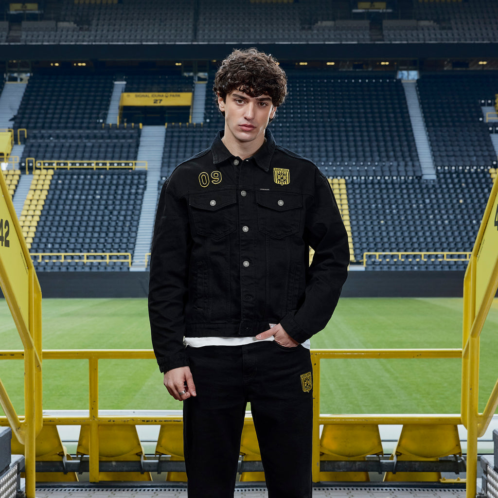 Black BVB x Guess denim jacket featuring the Borussia Dortmund logo and the number 09, modeled by a man at SIGNAL IDUNA PARK.