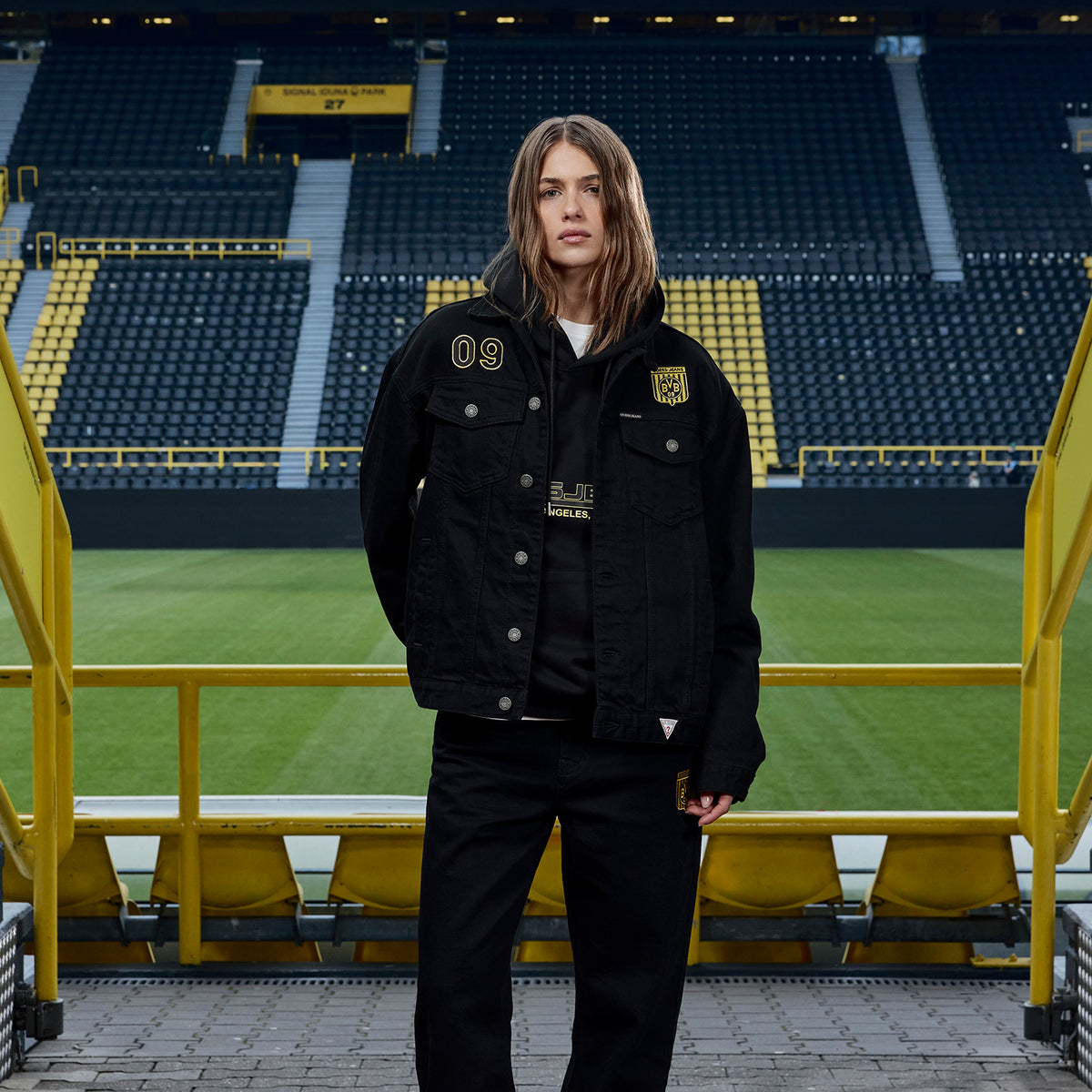 Schwarze BVB x Guess Jeansjacke mit gelbem BVB-Logo und der Zahl 09, getragen von einer Frau im SIGNAL IDUNA PARK.