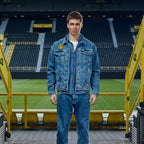 Veste en jean bleue BVB x Guess ornée de l'emblème du BVB, portée par un mannequin devant les tribunes du SIGNAL IDUNA PARK. Idéale pour les fans et les tenues stylées.