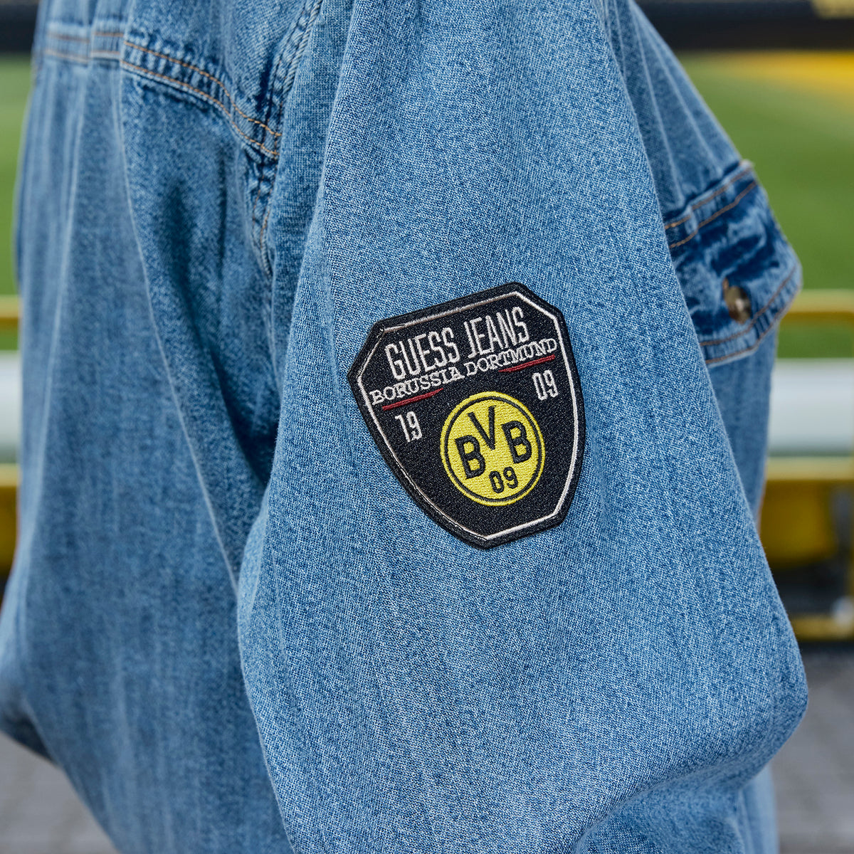BVB × Guess コレクションのブルーのデニム製ジーンズシャツ。袖にはBVBのロゴとGuess Jeansのパッチがあしらわれており、ボルシア・ドルトムントのファンに最適です。