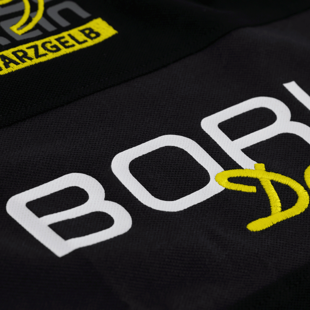 Vista detallada de un polo negro del BVB Elements II con el logotipo del BVB bordado en amarillo y blanco y la referencia al club.