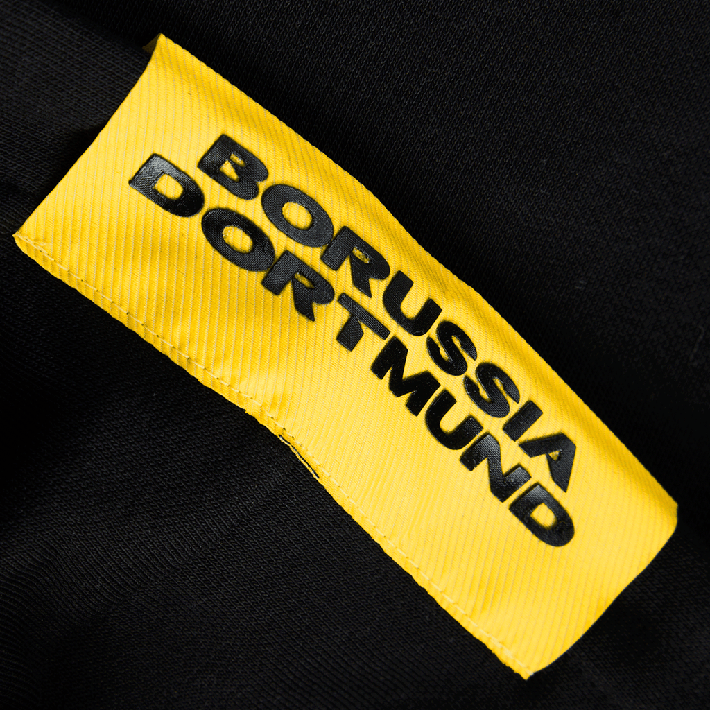 Detailansicht eines schwarzen BVB Hoodies der Essentials II Kollektion mit gelbem Borussia Dortmund Emblem. Idealer Fanartikel für Anhänger von Borussia Dortmund.