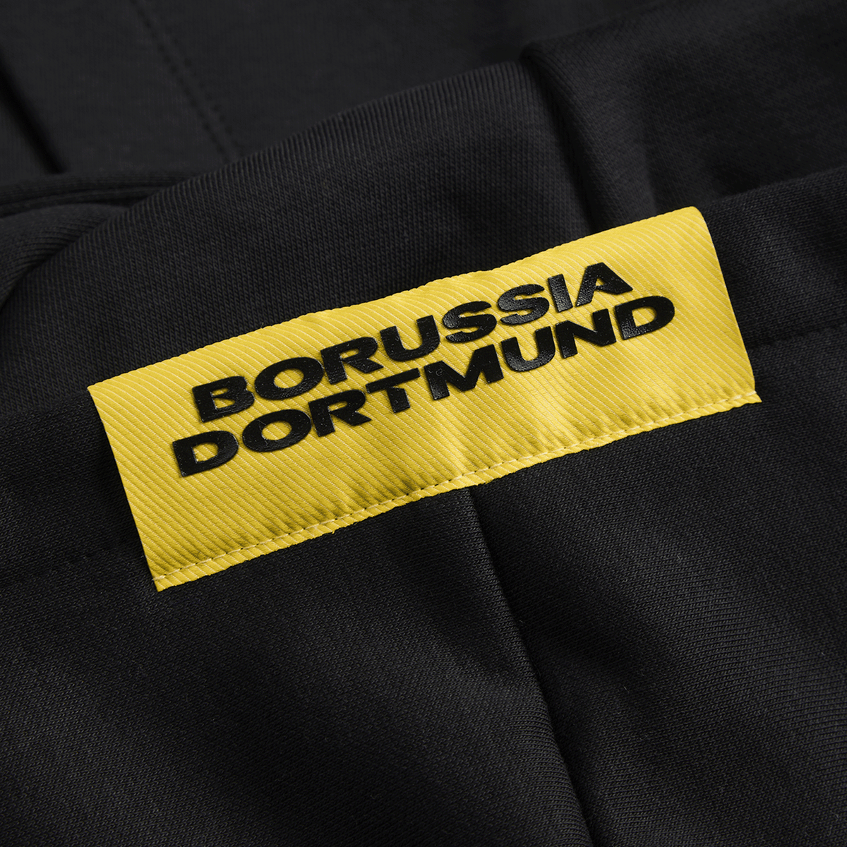 Immagine ravvicinata della giacca in felpa nera BVB Essentials II con etichetta gialla del Borussia Dortmund.