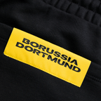 Gros plan sur le pantalon de survêtement noir BVB Essentials II, orné d'une étiquette jaune du Borussia Dortmund, idéal pour les supporters et les activités sportives.