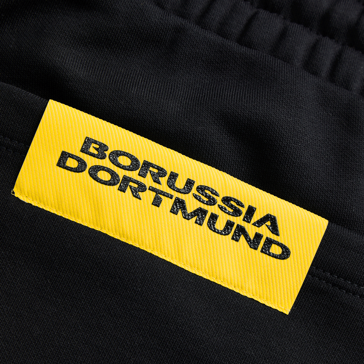 Gros plan sur le pantalon de survêtement noir BVB Essentials II, orné d'une étiquette jaune du Borussia Dortmund, idéal pour les supporters et les activités sportives.
